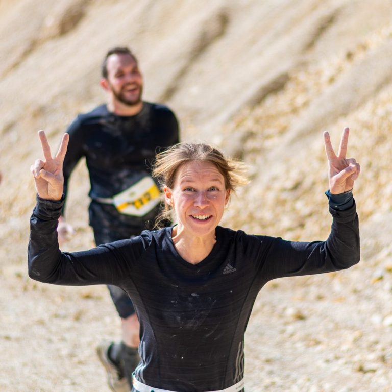 Am Ziel Muddy Älbler Läuferin mit Peace-Zeichen, begleitet von einem Mann im Hintergrund, auf unebenem Terrain.