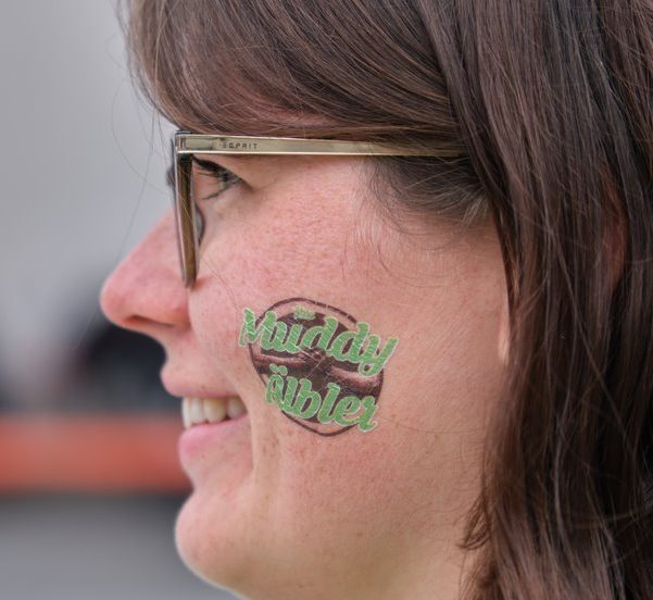 Frau mit einerMuddy Älbler logo Frau mit einerMuddy Älbler logo Gesichtsmarkierung und Brille lächelt seitlich.